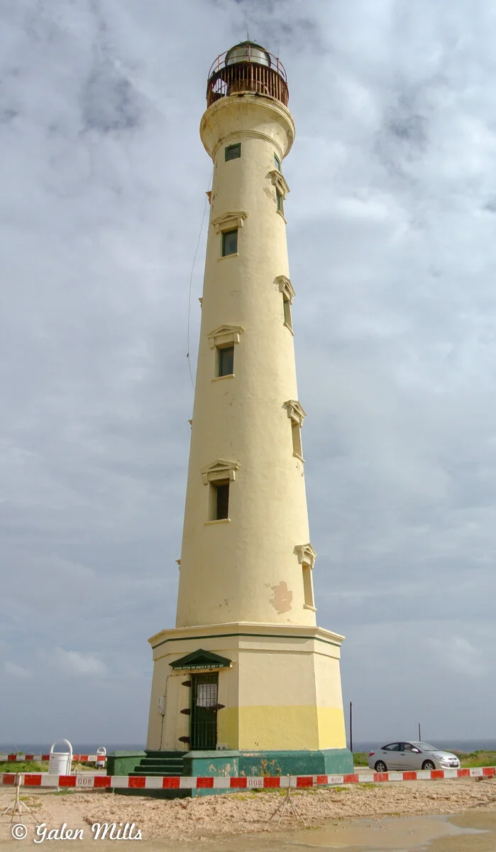 Tall yellow lighthouse with multiple windows, surrounded by red and white barriers, under a cloudy sky.