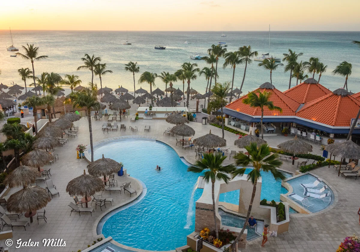 Aerial view of a tropical beach resort at sunset, featuring a large swimming pool, palm trees, thatched umbrellas, and a clear ocean with boats.