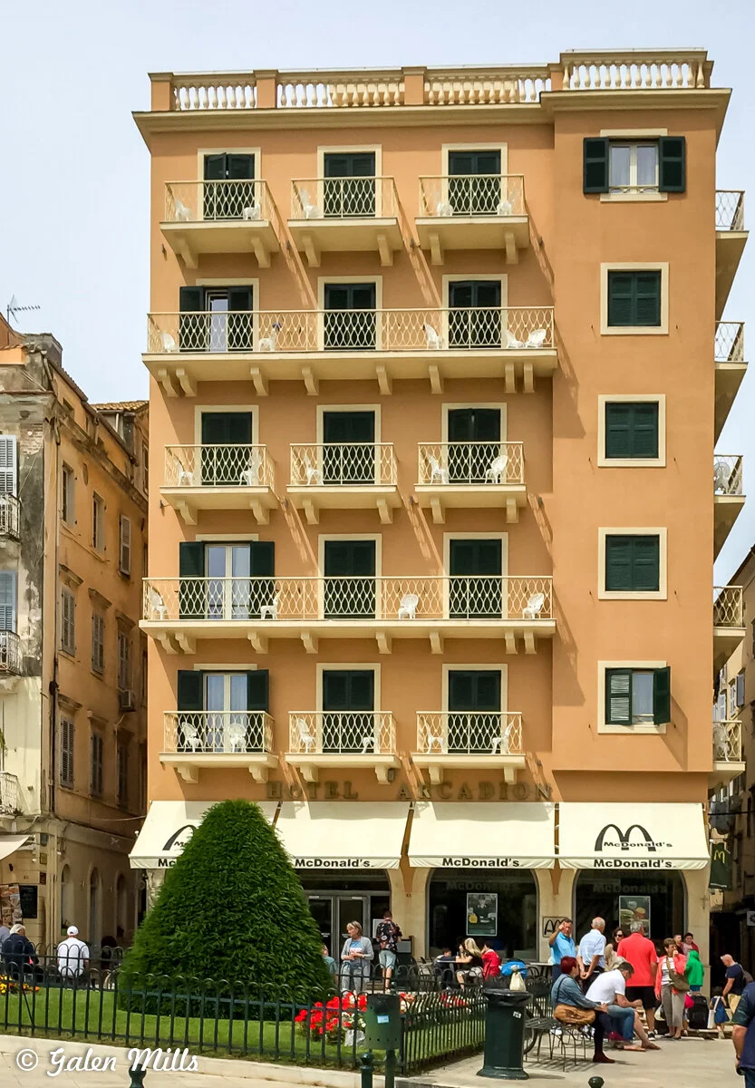 Beige multi-story building with balconies, green shutters, a McDonald's on the ground floor, and people sitting outside, with a manicured lawn and plants in front.