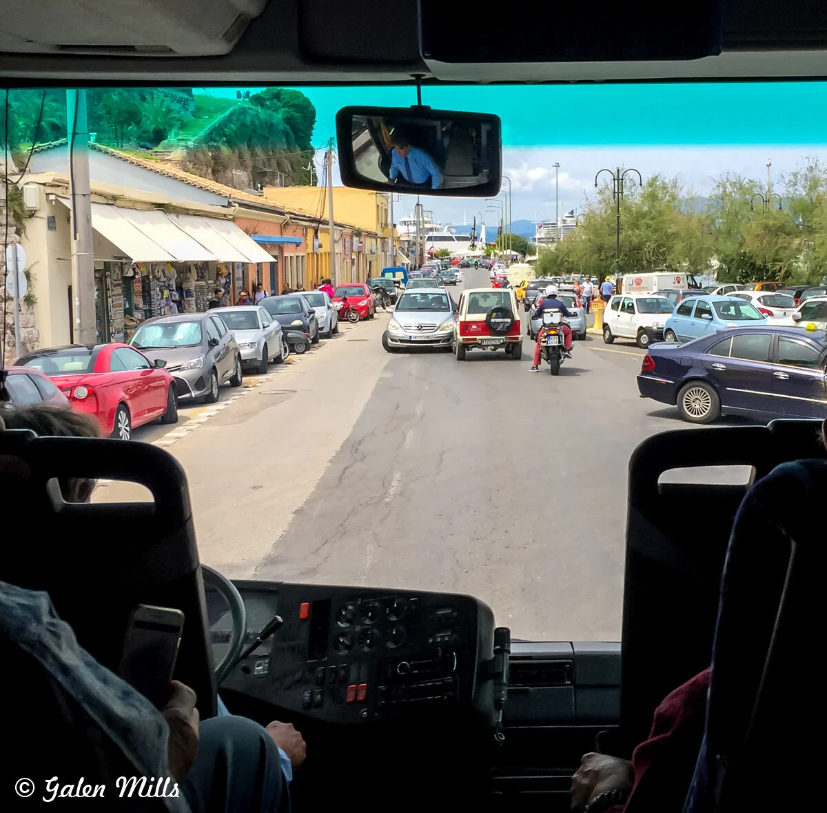View from inside a bus on a busy street with parked cars and a motorcycle. Shops with awnings line the left side.