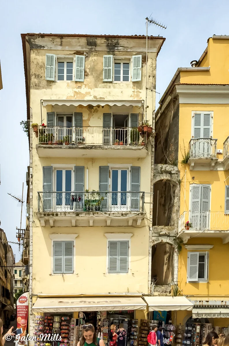 Old building with weathered facade, green shutters, balconies with plants, and a souvenir shop below in Corfu, Greece.