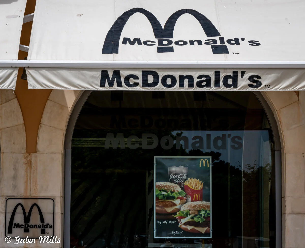 McDonald's restaurant entrance with white awning displaying the McDonald's logo, glass door featuring a promotional poster for a meal deal with Coca-Cola Zero, fries, and burgers.