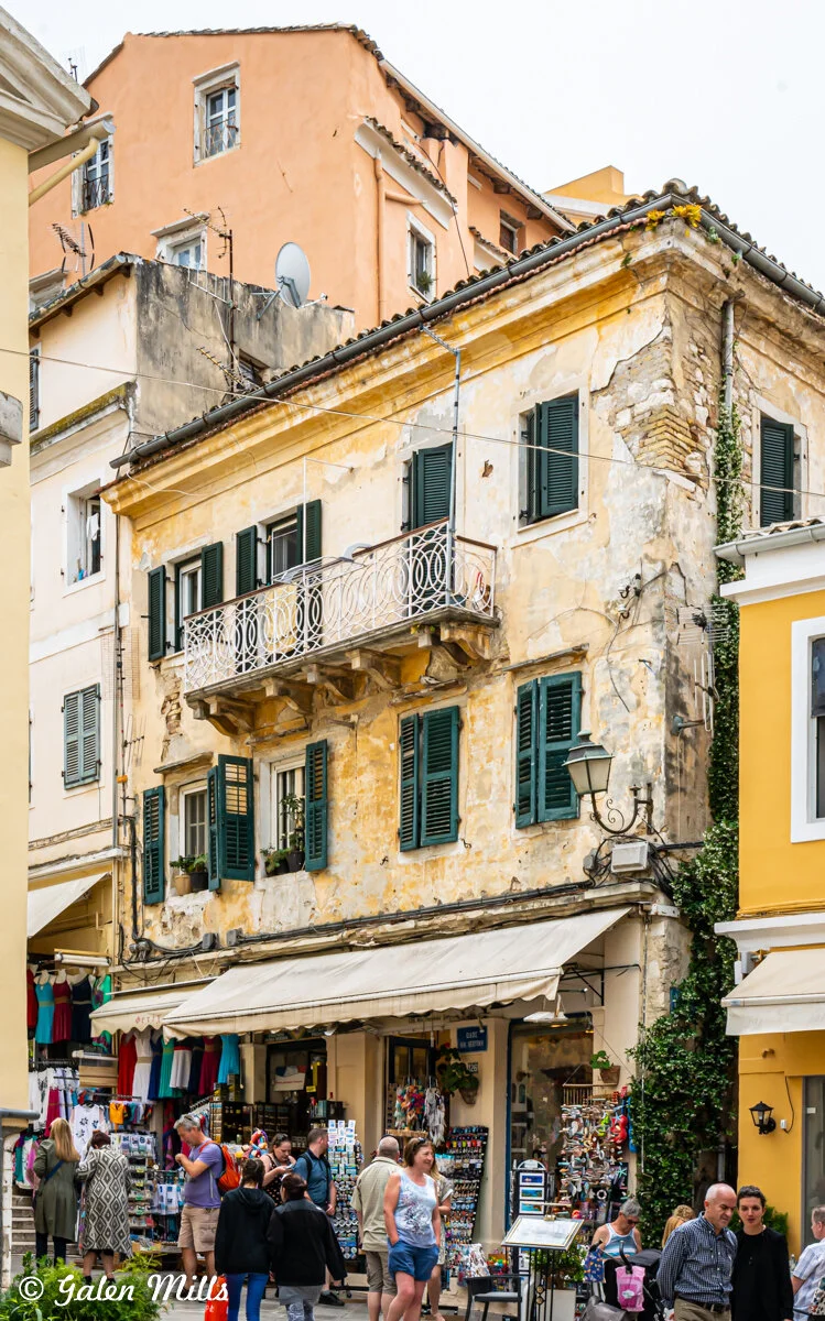 Street scene with an old building featuring yellow peeling paint and green shutters, a white iron balcony, and shops below. People are casually shopping and walking.