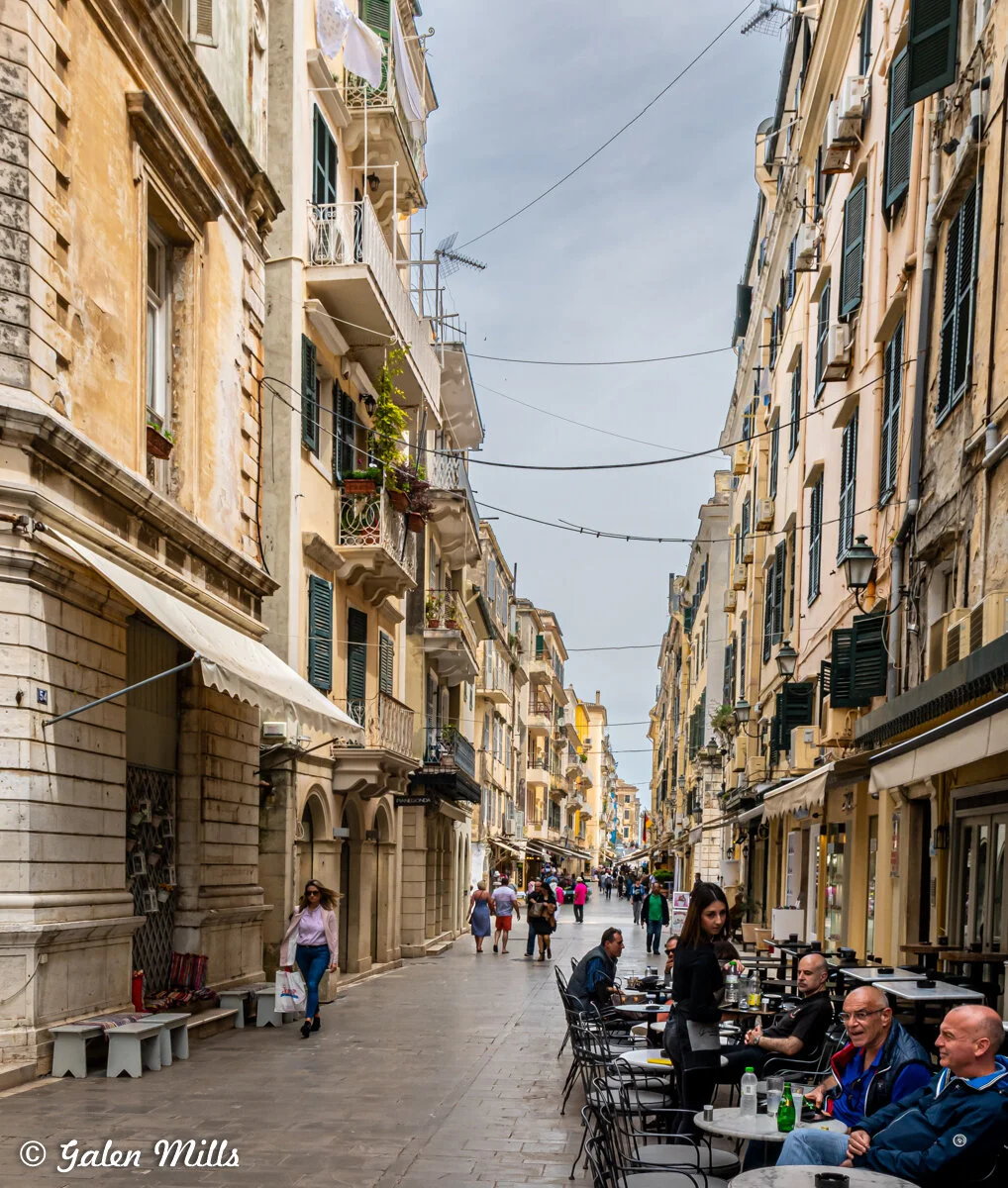 Narrow European street lined with historic buildings, outdoor cafes, and pedestrians on a cloudy day.