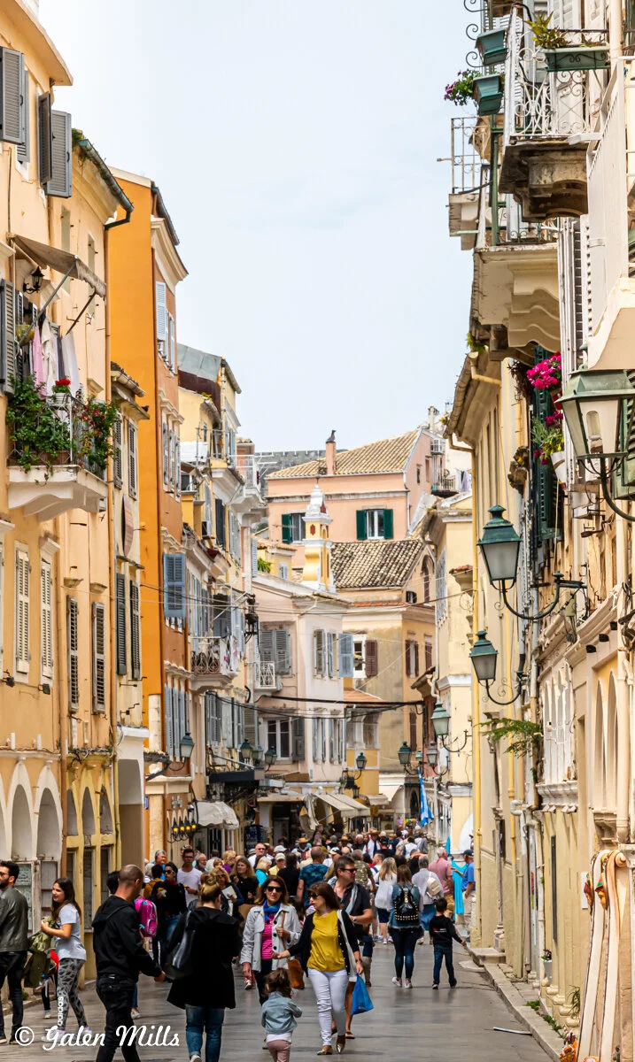 Busy European street scene with colorful buildings, balconies, flower pots, and people walking.