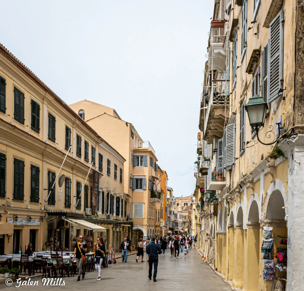 A pedestrian street lined with historic buildings featuring shutters and balconies; people walking and a few seated at outdoor cafe tables.