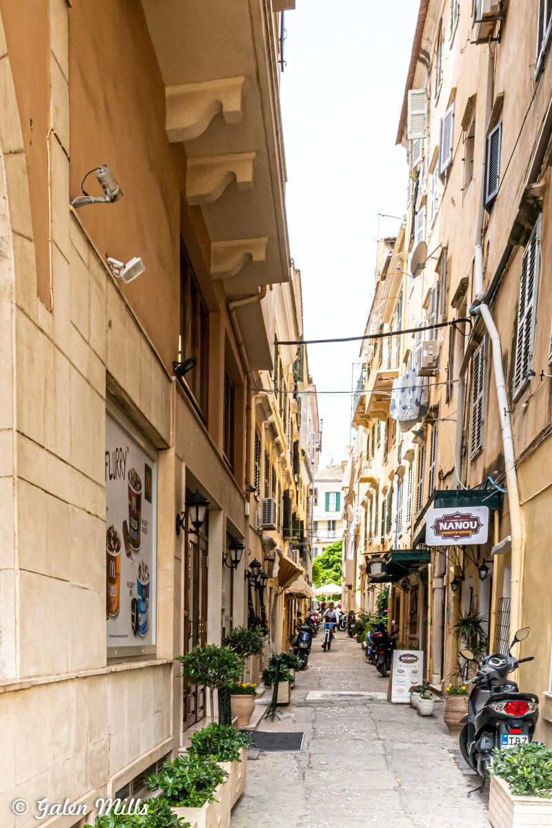 Narrow European street with shops, plants, and motorbikes. Signs for cafes and restaurants line the buildings. People can be seen in the distance.