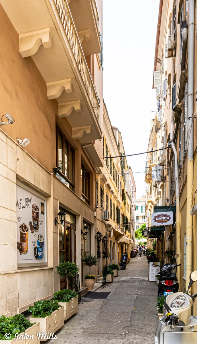 Narrow European street with tall buildings, wall advertisements, and a store named 'Nanou'; scooters parked alongside.