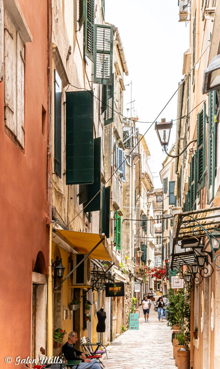 Narrow European street lined with colorful buildings with green shutters, hanging clotheslines, outdoor seating, and flower pots. Wandering pedestrians and a mannequin outside a shop. "Art Forum" sign visible.