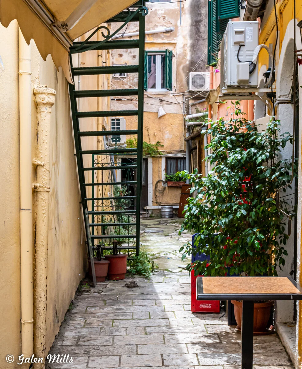 Narrow alleyway with old buildings, green plants, a staircase, and an outdoor table.