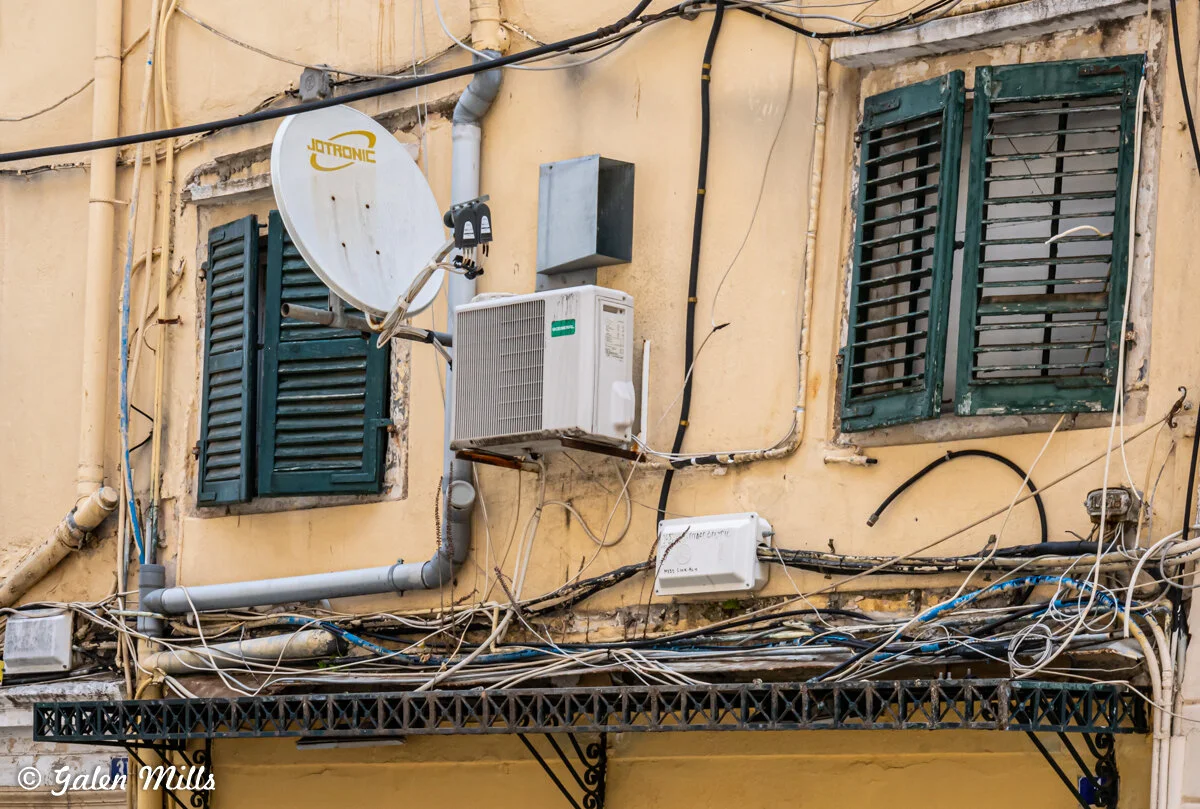Close-up of a building facade with a satellite dish, air conditioning unit, and tangled wires near green shutters on a yellow wall.