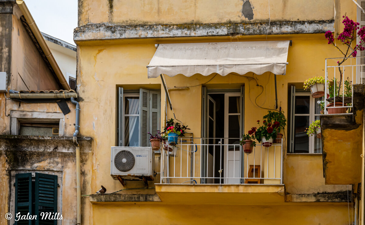 A small balcony with white railing on a yellow building exterior. The balcony is decorated with potted plants and flowers, and there's an air conditioning unit mounted on the wall. An awning is extended above the balcony door. A pigeon is perched nea