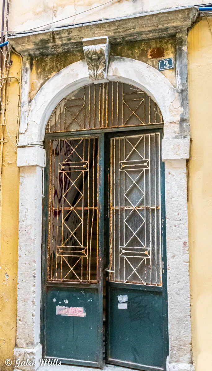 Old metal gate with ornate design and archway, weathered stone frame, and a number plaque "3" on a yellow wall.