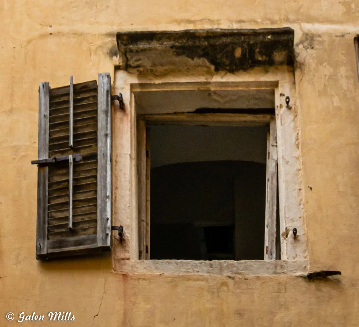 Old weathered window with one open wooden shutter on a distressed, yellow-painted wall.