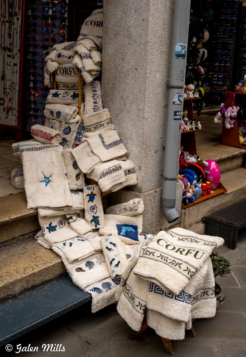 Assortment of decorative towels with Greek patterns and 'Corfu' text displayed outside a souvenir shop.