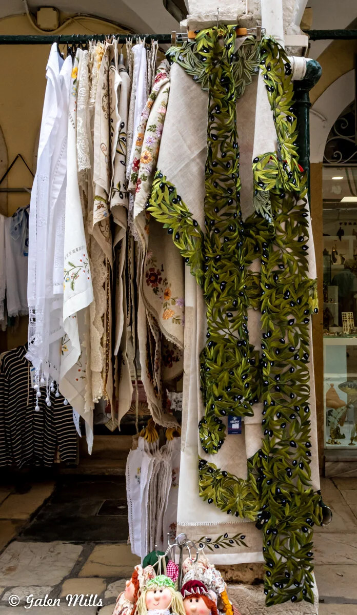Display of embroidered textiles and olive branch-patterned garments hanging at a market stall, with small decorative dolls at the bottom.