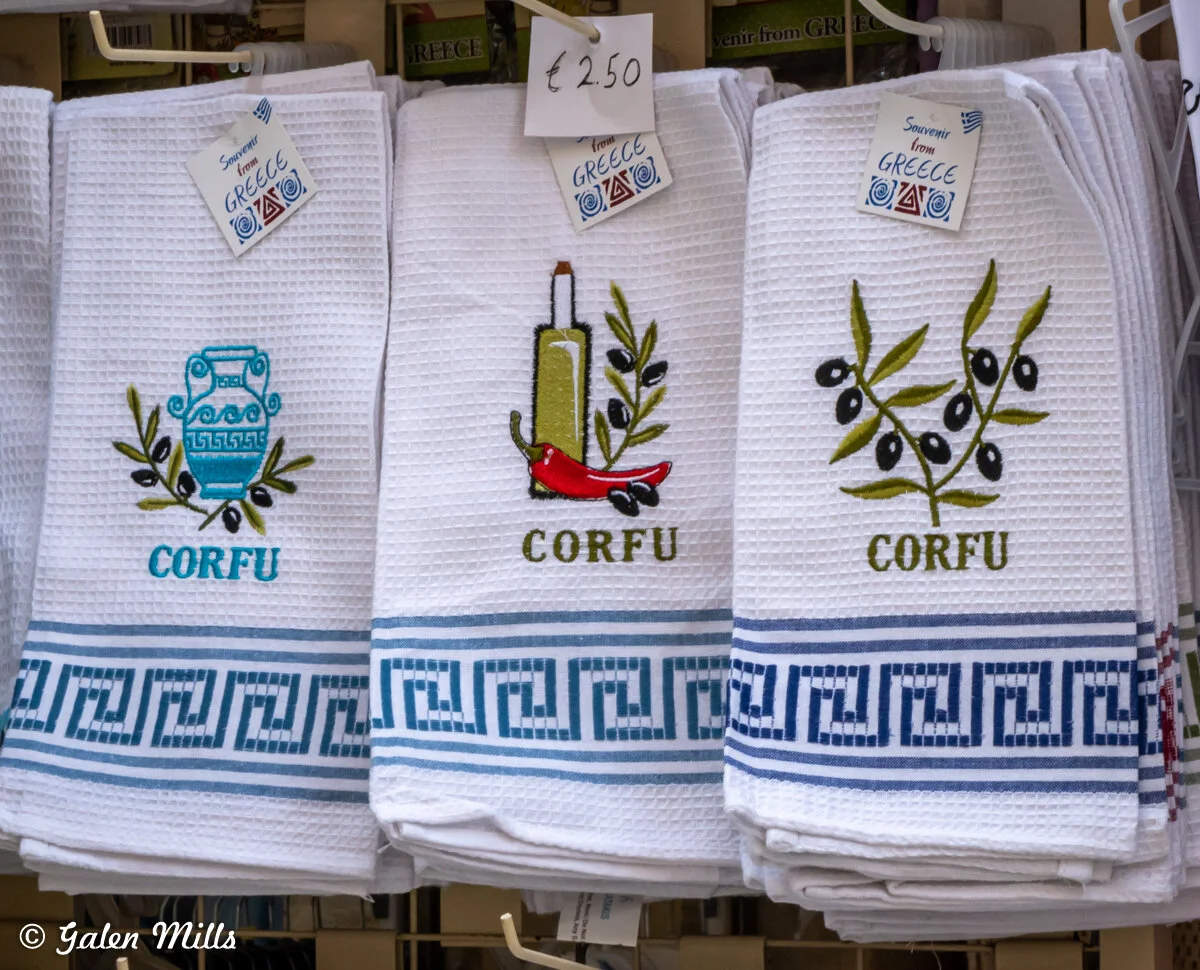 Greek souvenir towels from Corfu, featuring embroidered designs of a vase, olive oil bottle with a chili, and olive branch, priced at €2.50.