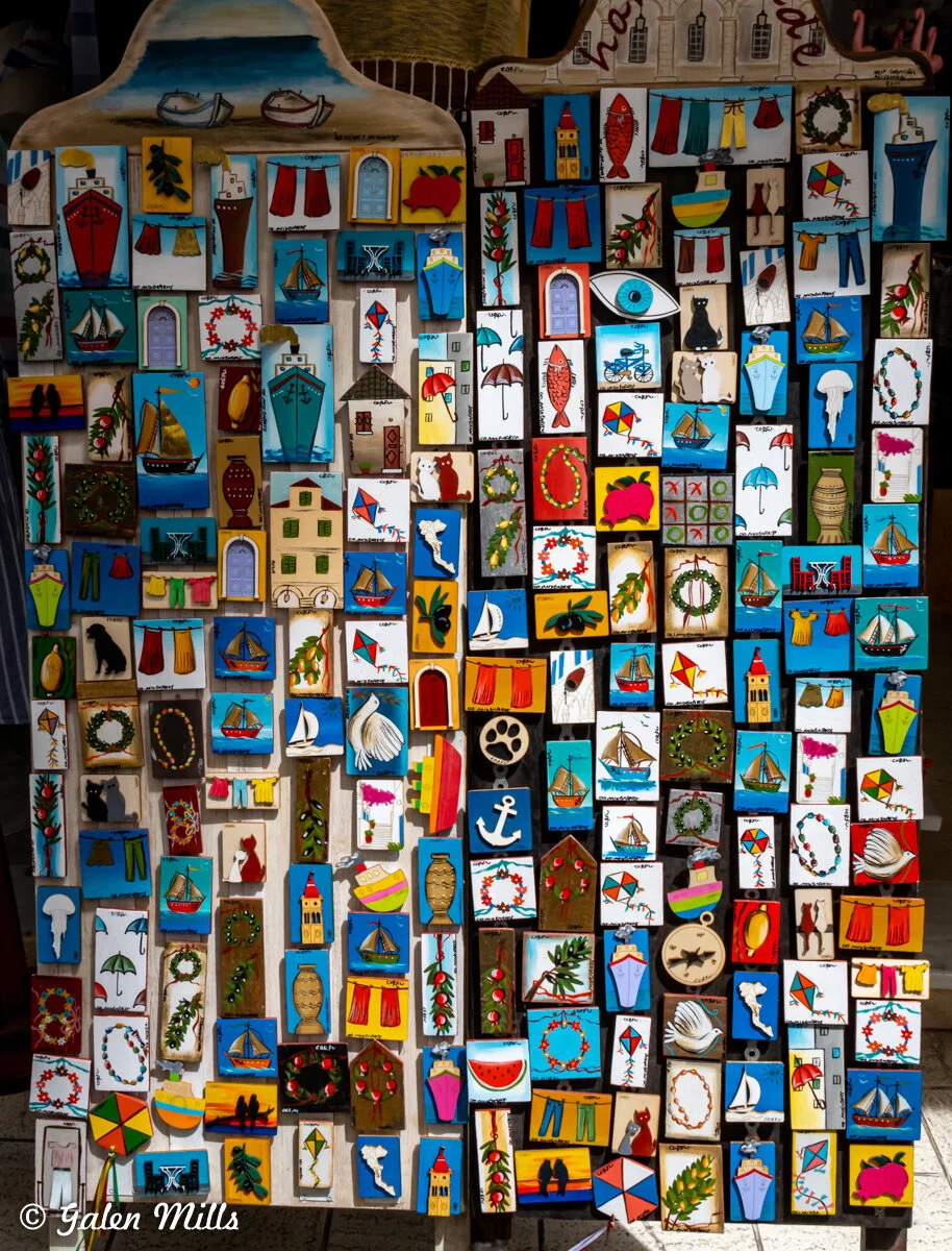 Display of colorful handmade magnets including nautical and seasonal designs such as boats, lighthouses, anchors, kites, and wreaths, on a wooden board.