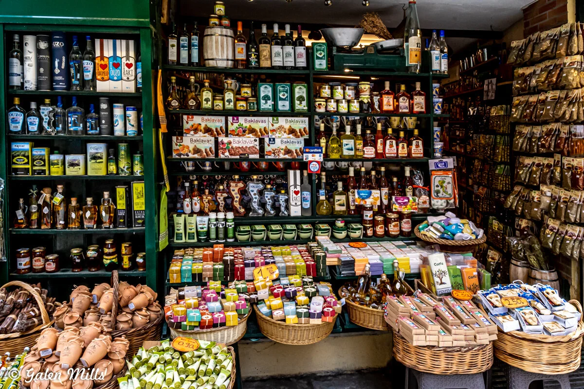 Market stall with shelves of jars, bottles, and packages of various goods, including olive oil, spices, and soaps. Baskets with traditional items and local delicacies are also displayed.