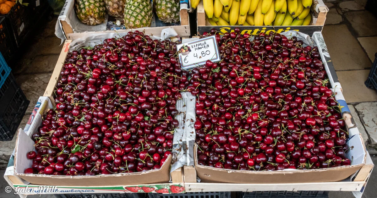 Boxes of cherries displayed at a street market with pineapples and bananas in the background.