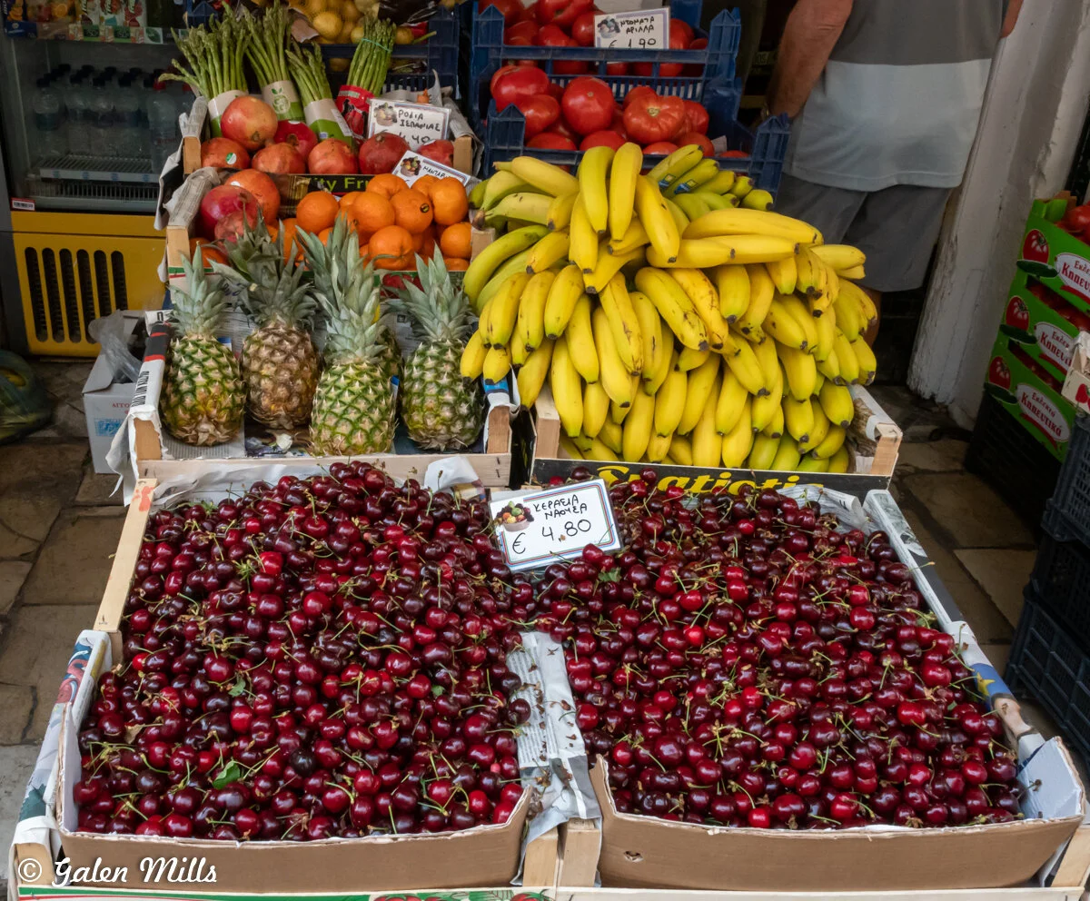 Market display of fresh fruits including cherries, bananas, pineapples, oranges, pomegranates, and tomatoes.