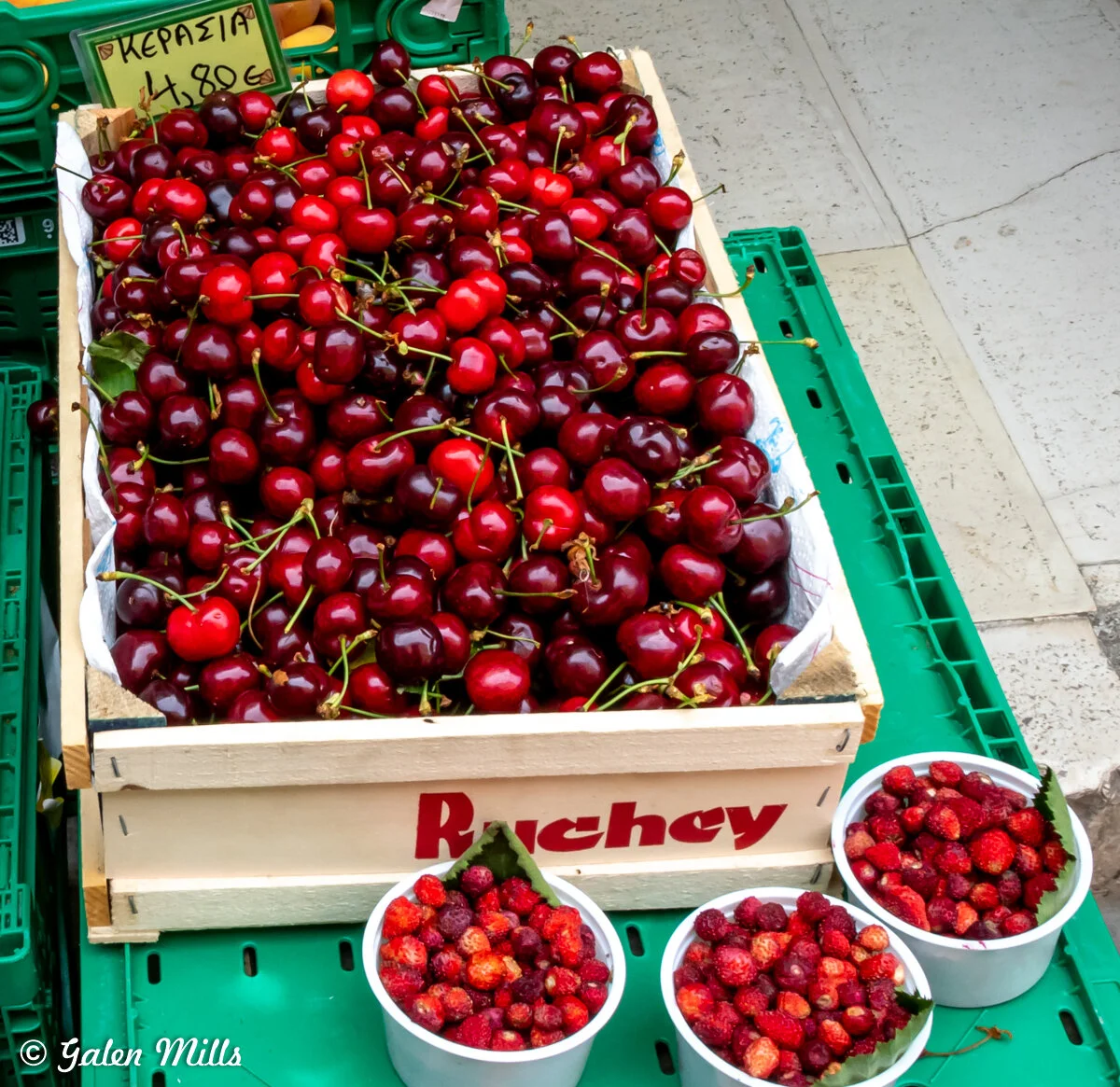 A wooden crate filled with fresh red cherries, surrounded by three small containers of wild strawberries, placed on a green market stand.