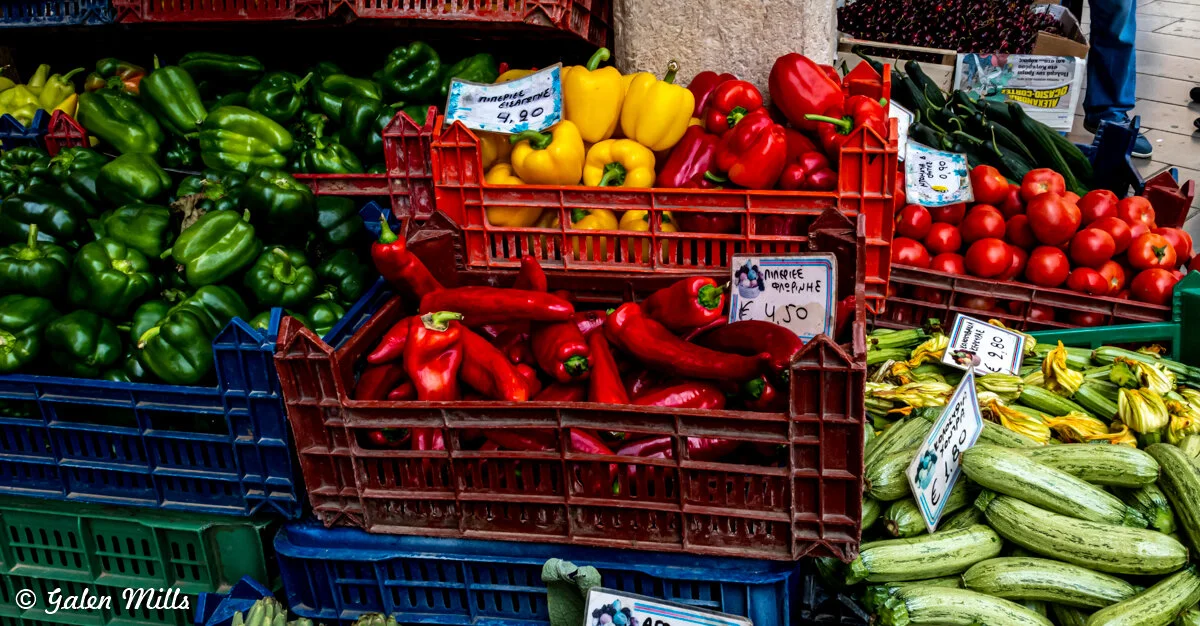 Assorted colorful vegetables including bell peppers, tomatoes, zucchini, and cucumbers displayed in baskets at a market.
