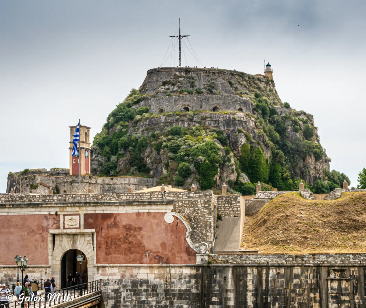 The Old Fortress of Corfu in Greece under a cloudy sky, with historical buildings and a Greek flag visible.