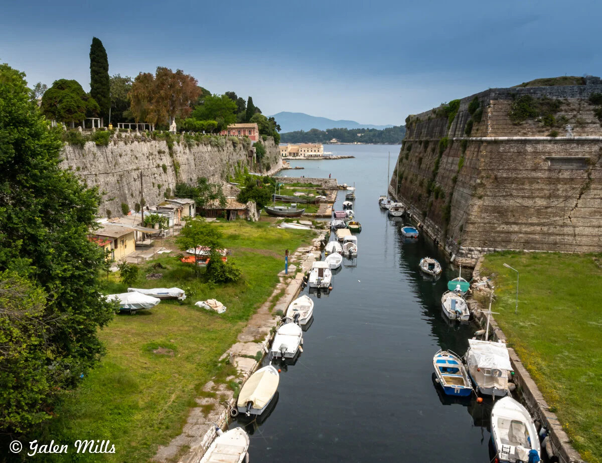 Narrow canal lined with small boats next to historic stone walls and green grassy banks. Dense tree area and old buildings visible beyond the canal. Overcast sky in the background.