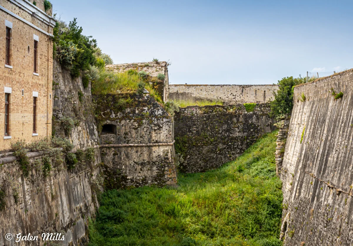 Ancient stone fort walls with greenery, featuring a brick building on the left and overgrown vegetation in the moat area.