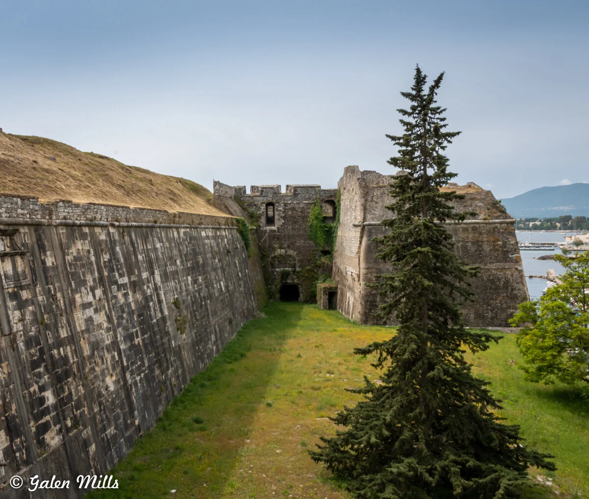 Old fortress with stone walls, grassy area, and a tall tree, overlooking a body of water and distant mountains.