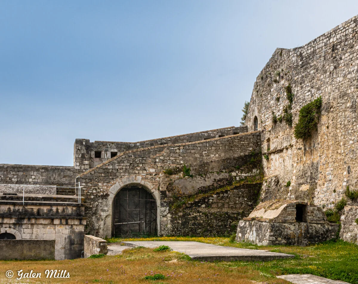 Old stone fortress with arched entrance, weathered walls, and patches of greenery against a clear blue sky.