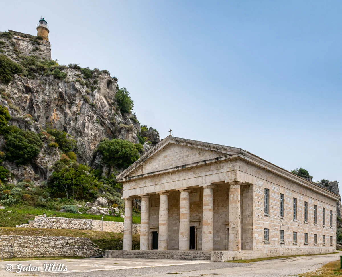 A neoclassical church with columns and a gabled roof is in the foreground. Behind it is a rocky cliff with a lighthouse on top. The sky is clear and blue.