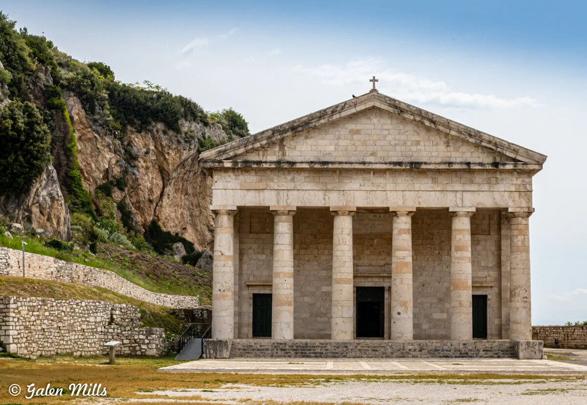 Stone building with classical columns and pediment, resembling a Greek temple, set against a rocky hillside and green vegetation.