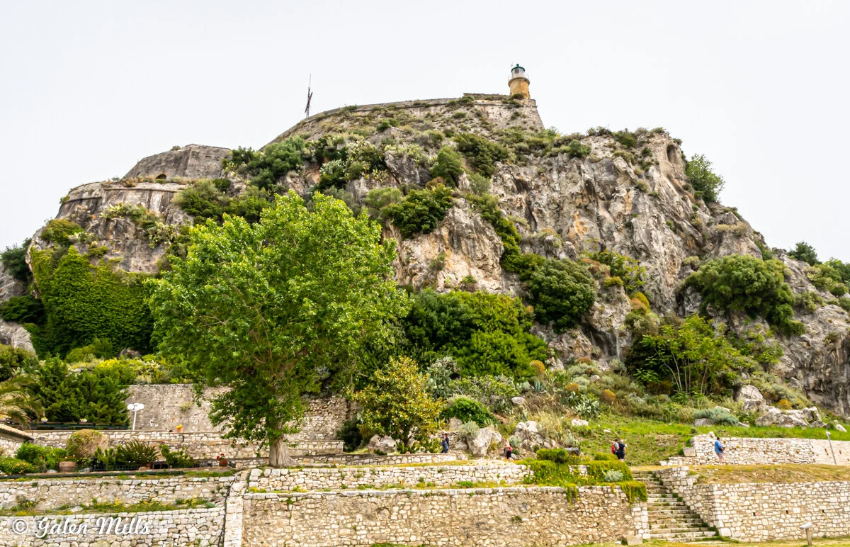 Rocky hill with lush greenery and a stone walkway, featuring a lighthouse at the top.