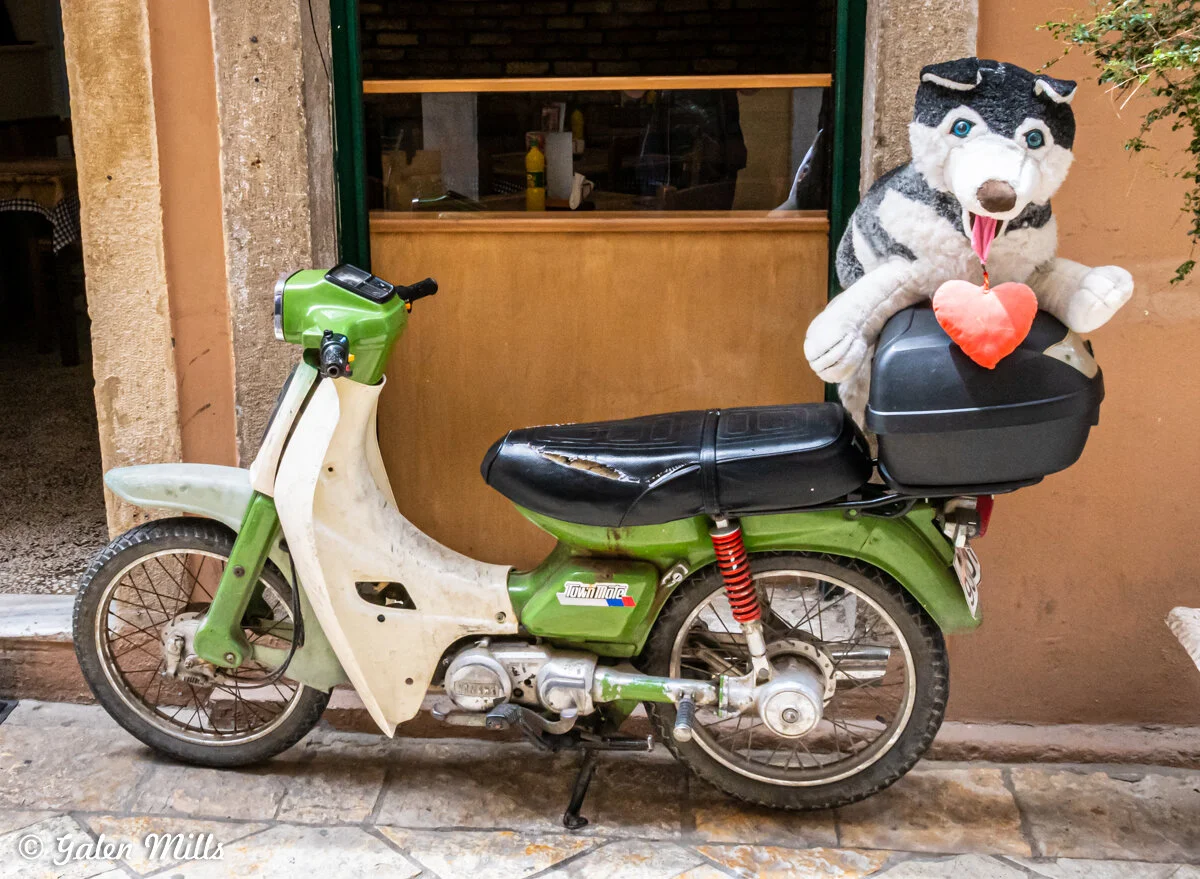 Green and white vintage moped with a plush dog toy holding a red heart on the back seat, parked in front of a building.