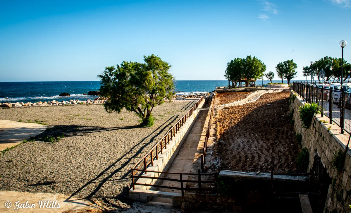 Coastal landscape with trees, a sandy path, and a rocky shoreline, against a backdrop of the ocean and blue sky.
