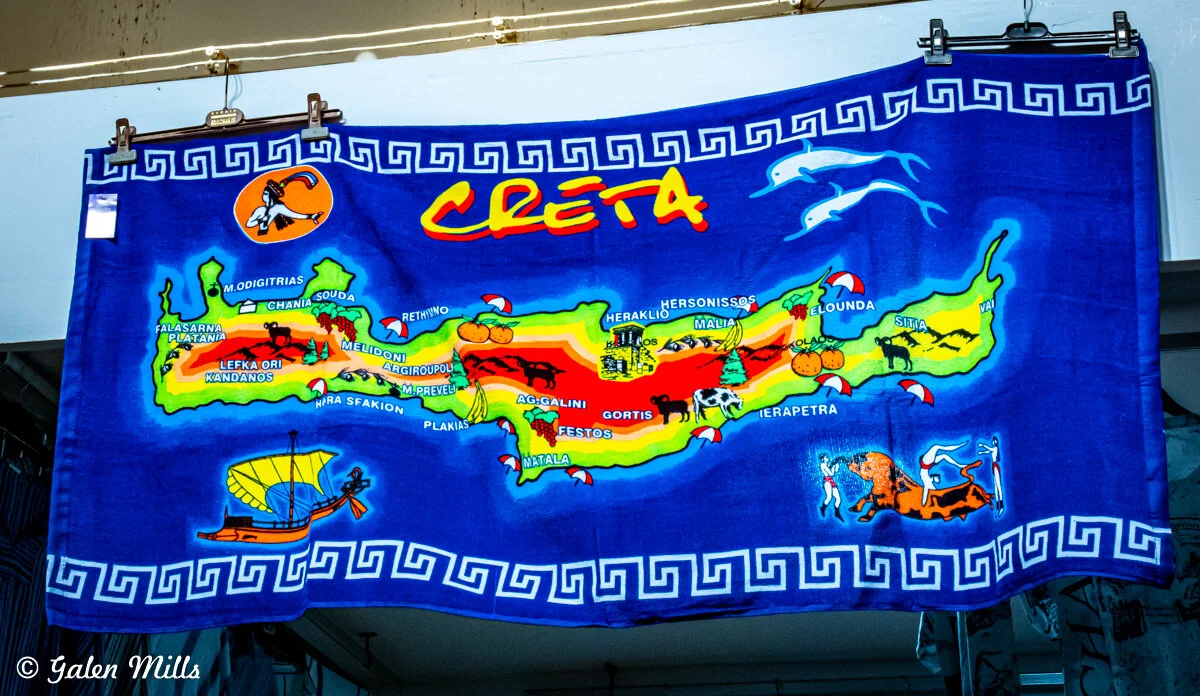 Colorful towel featuring a map of Crete with illustrations of landmarks, animals, and regional names on a blue background.