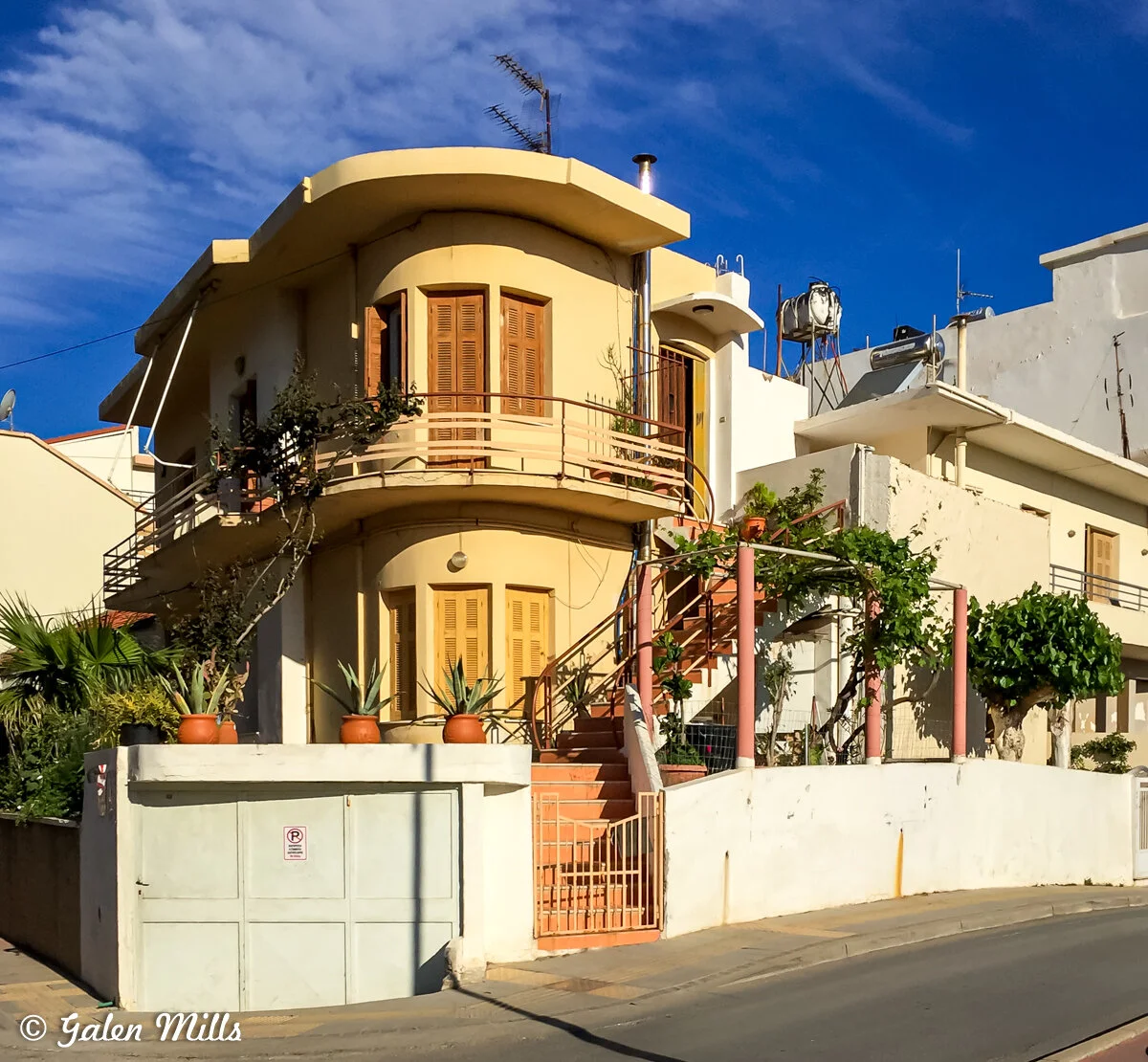Two-story cream-colored house with curved balcony, wooden shutters, and potted plants; exterior staircase; white walls and clear sky.