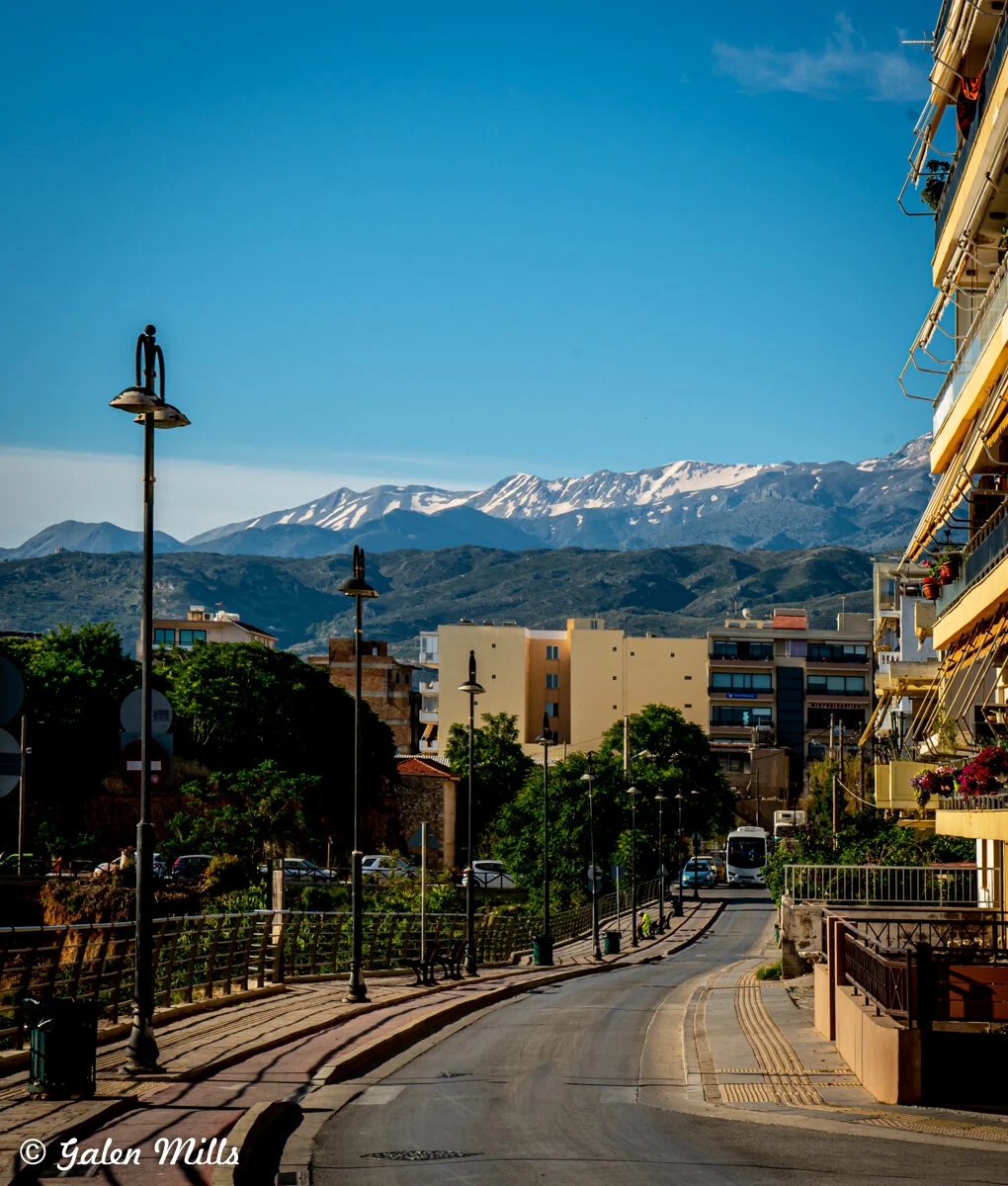 City street with modern buildings, street lamps, and distant snow-capped mountains under clear blue sky.