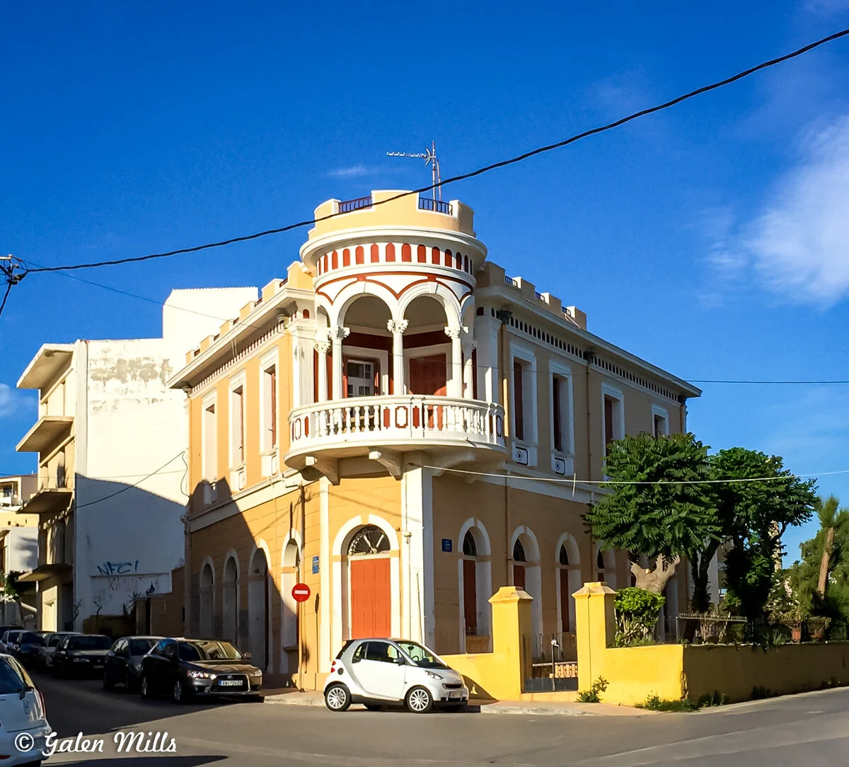 Yellow and white neoclassical building with a round balcony, parked cars, and blue sky.