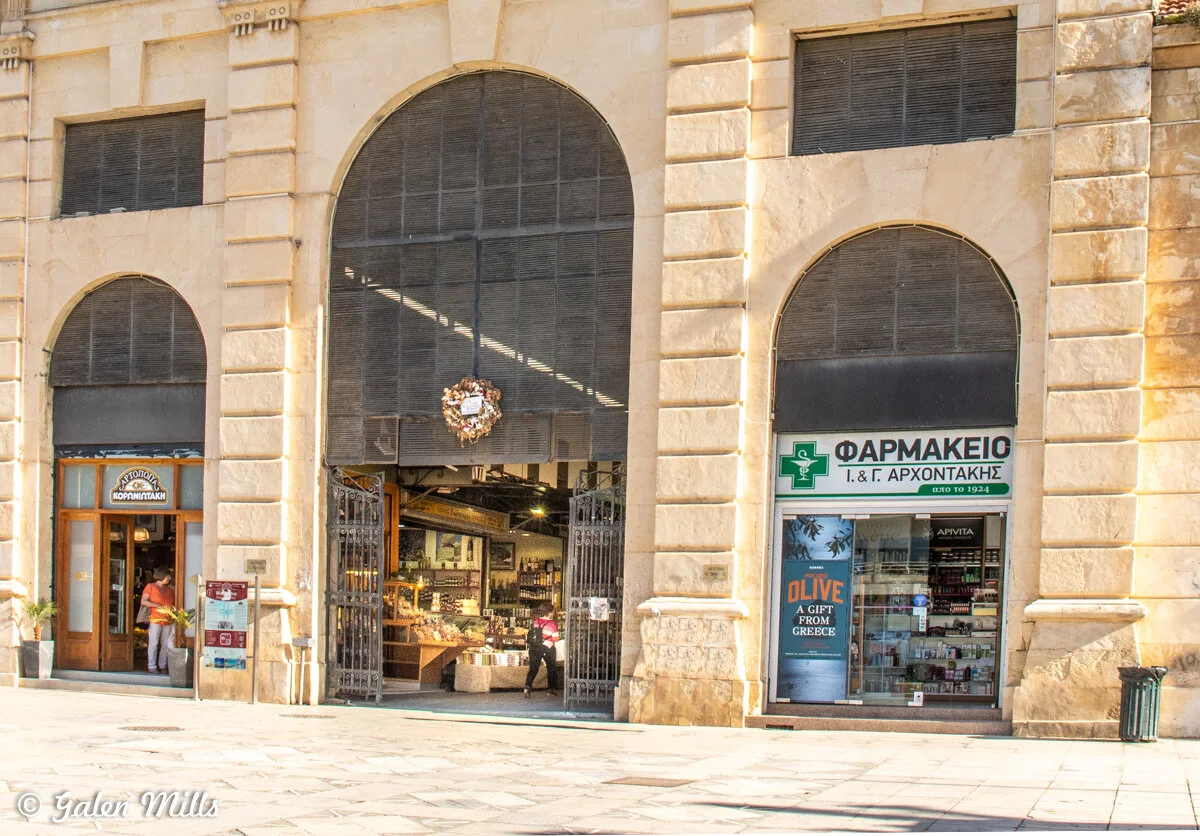 Exterior of historic building with a pharmacy and shop entrance in Heraklion, Crete. The pharmacy sign is in Greek, offering products like olive oil as gifts from Greece. The facade features large arches and stonework.