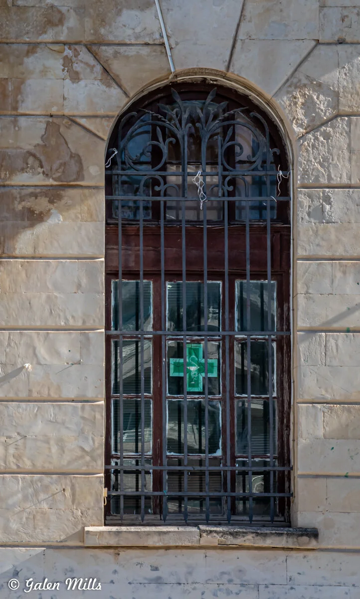 Vintage arched window with iron bars and green cross sign on weathered stone wall.