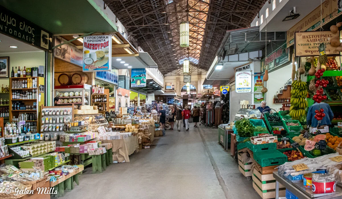 Indoor market with various stalls selling goods such as fruits, vegetables, cheeses, and packaged products. People are shopping and walking through the market. Overhead lighting and signs with Greek text are visible.