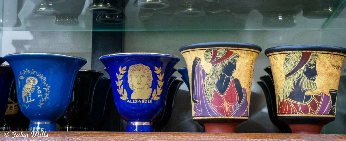 Collection of decorative ancient Greek-style pottery cups, featuring blue and black designs with gold accents, including figures of an owl, Alexander the Great, and Greek mythological characters.