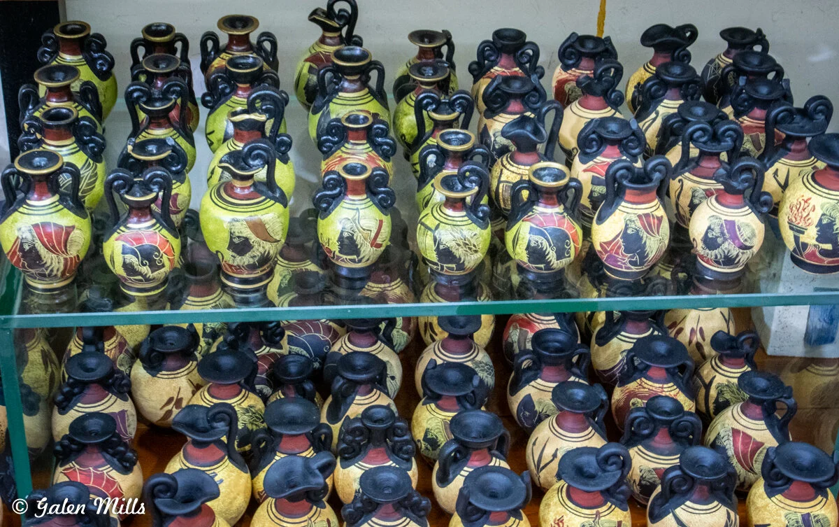 Collection of small, ancient Greek-style amphorae with black and red figures, displayed on glass shelves.