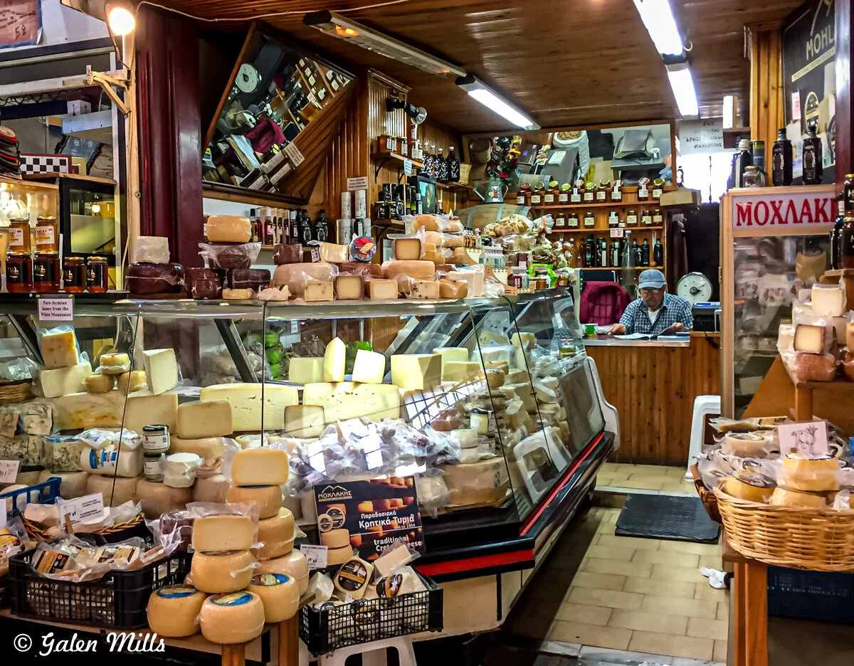 Cheese shop interior with various cheeses displayed behind a glass counter. Assorted bottles and jars are on shelves in the background. A person is seated at a counter, working with documents.