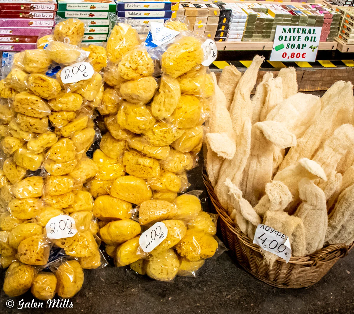 Sponges in plastic bags, natural olive oil soap, and loofahs in a market display.