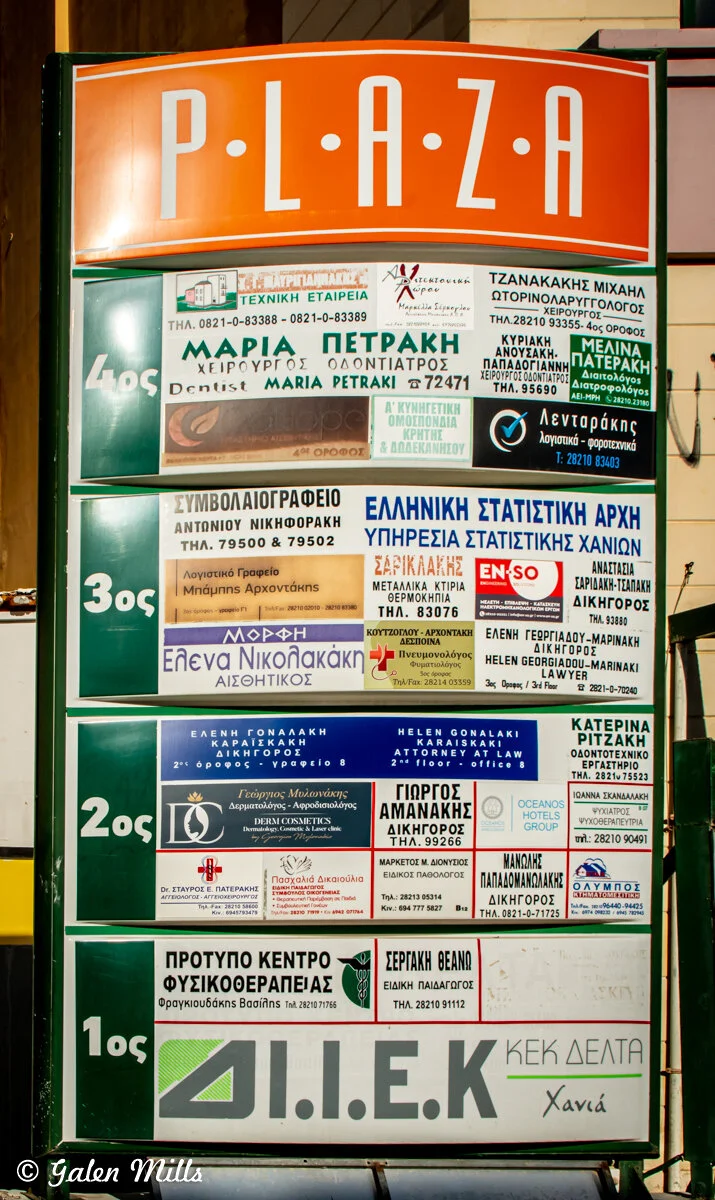 Directory sign for Plaza building, displaying names and contact information for various businesses, primarily in Greek, including dentists, lawyers, and other professional services. Sign is divided by floors with an orange top header reading 'PLAZA.'