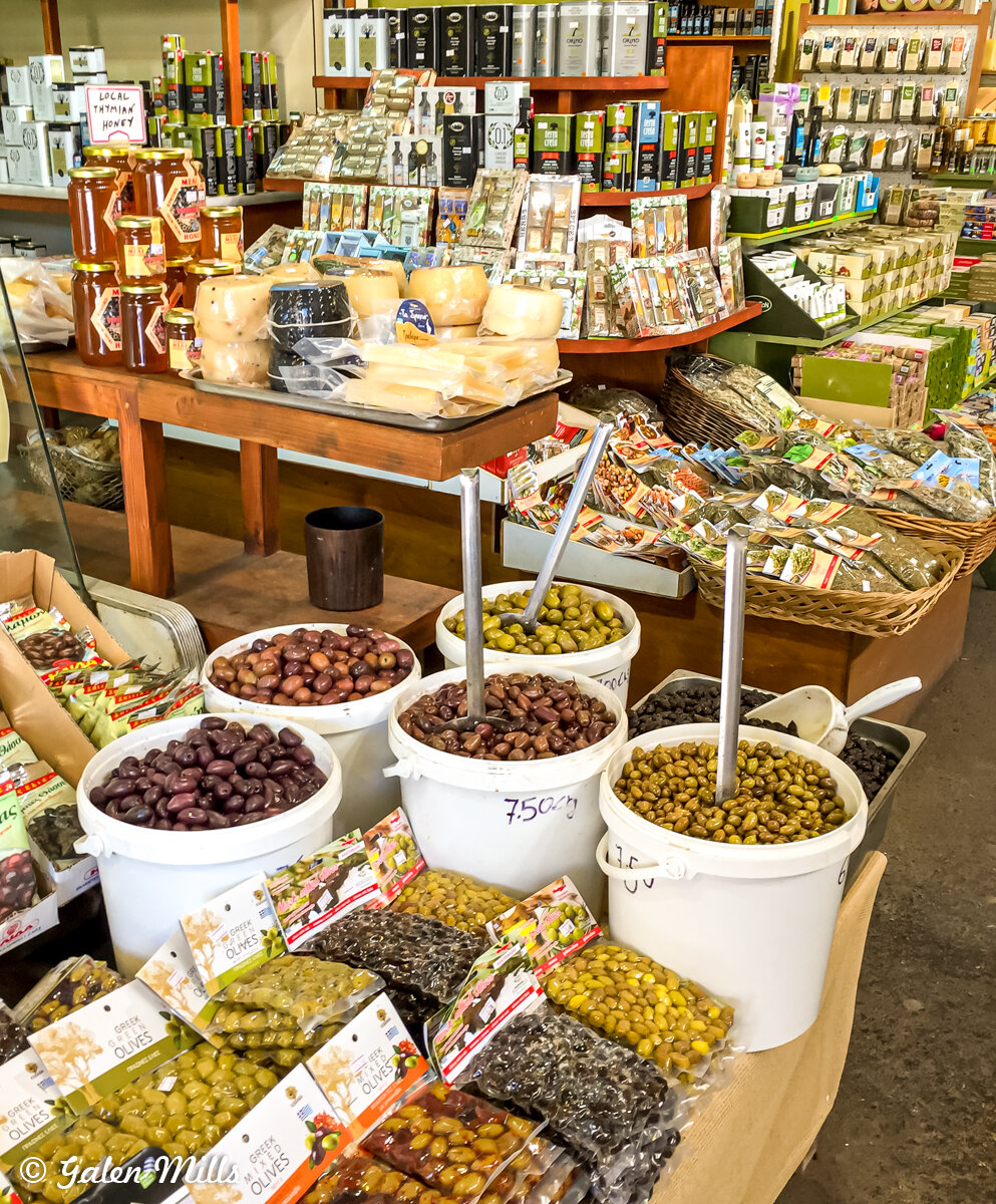 Market display with buckets of various olives, packaged goods, jars of honey, and cheese wheels on a wooden table.